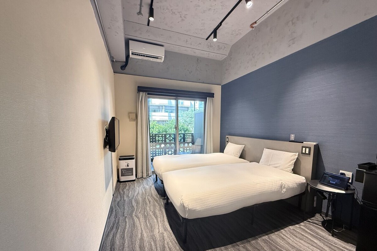 A modern hotel room with a large bed, a desk with a laptop, and a view of greenery outside.