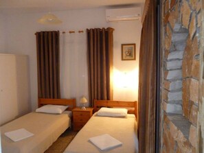 A hotel room with two beds, a nightstand, an air conditioning unit, and a stone wall. - Vasilis Ios Place Room 3 (Chora)
