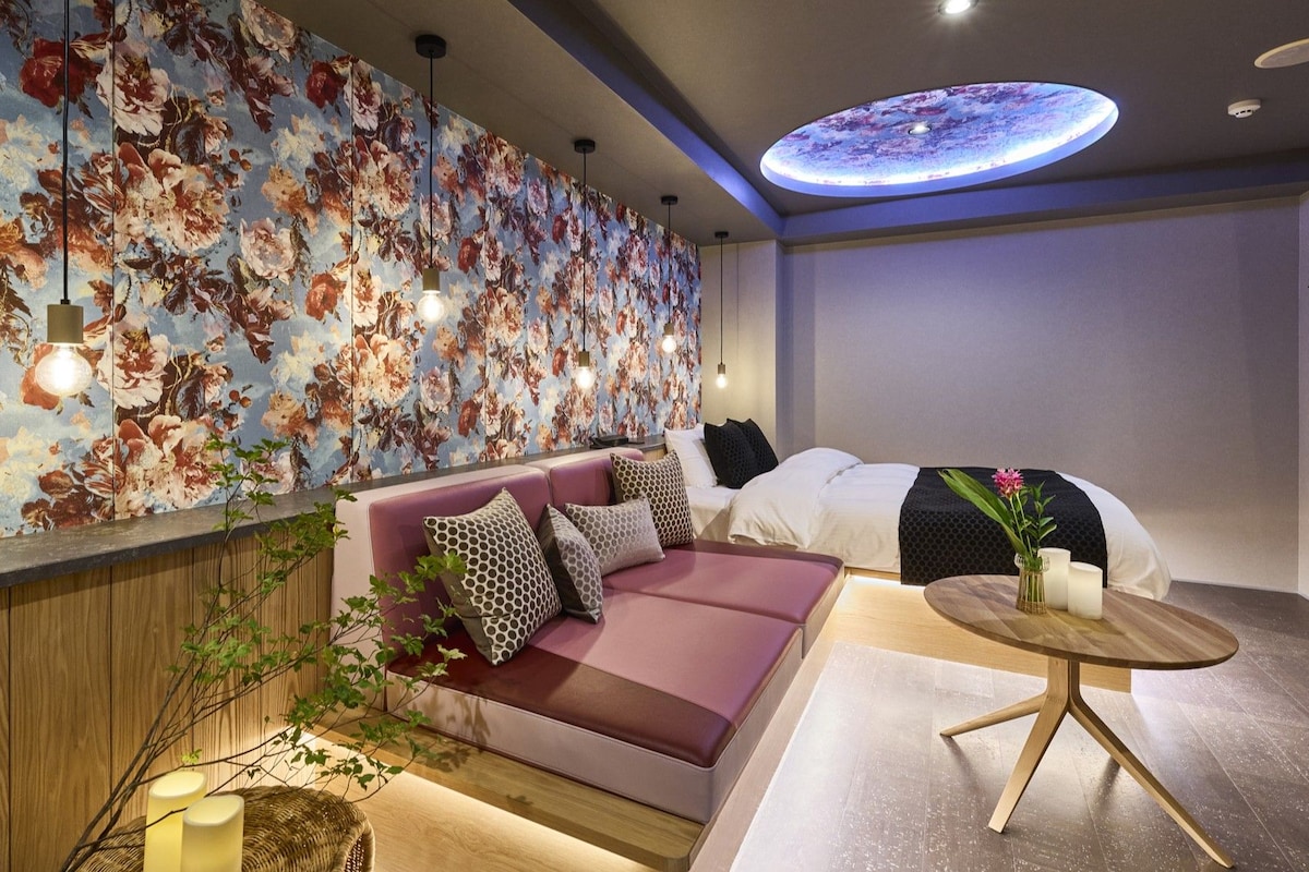 A modern hotel room with a floral wallpaper, a sofa, a bed, a round wooden table, and a ceiling light.