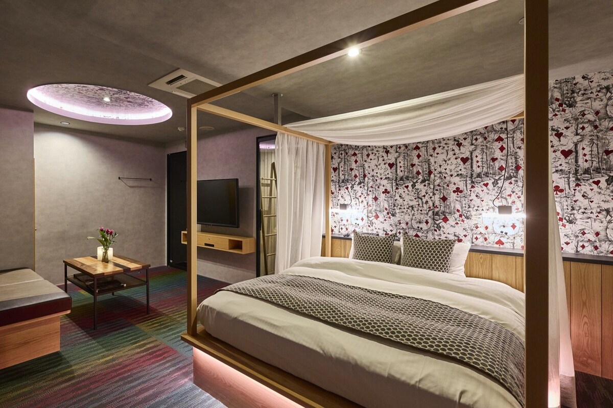 A modern hotel room with a large bed, a television, a small table with a vase, and a colorful carpet.