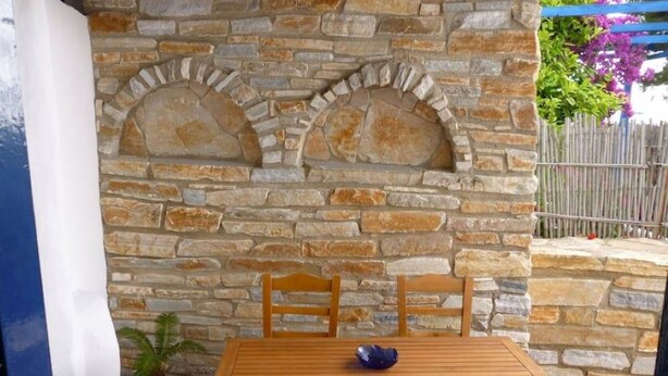 A stone wall with a wooden table and chairs, a potted plant, and a blue railing. - Vasilis Ios Place Room 1 (Chora)