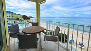 Ein Balkon mit rundem Tisch und vier Stühlen, mit Blick auf einen Strand und einen Poolbereich.
