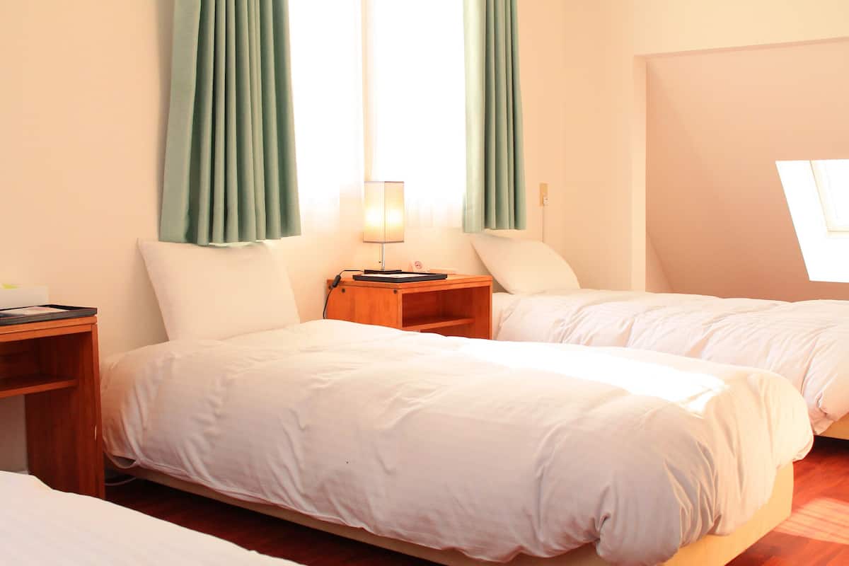 Quarto de hotel com duas camas, uma mesa de cabeceira com abajur e uma janela com cortinas.