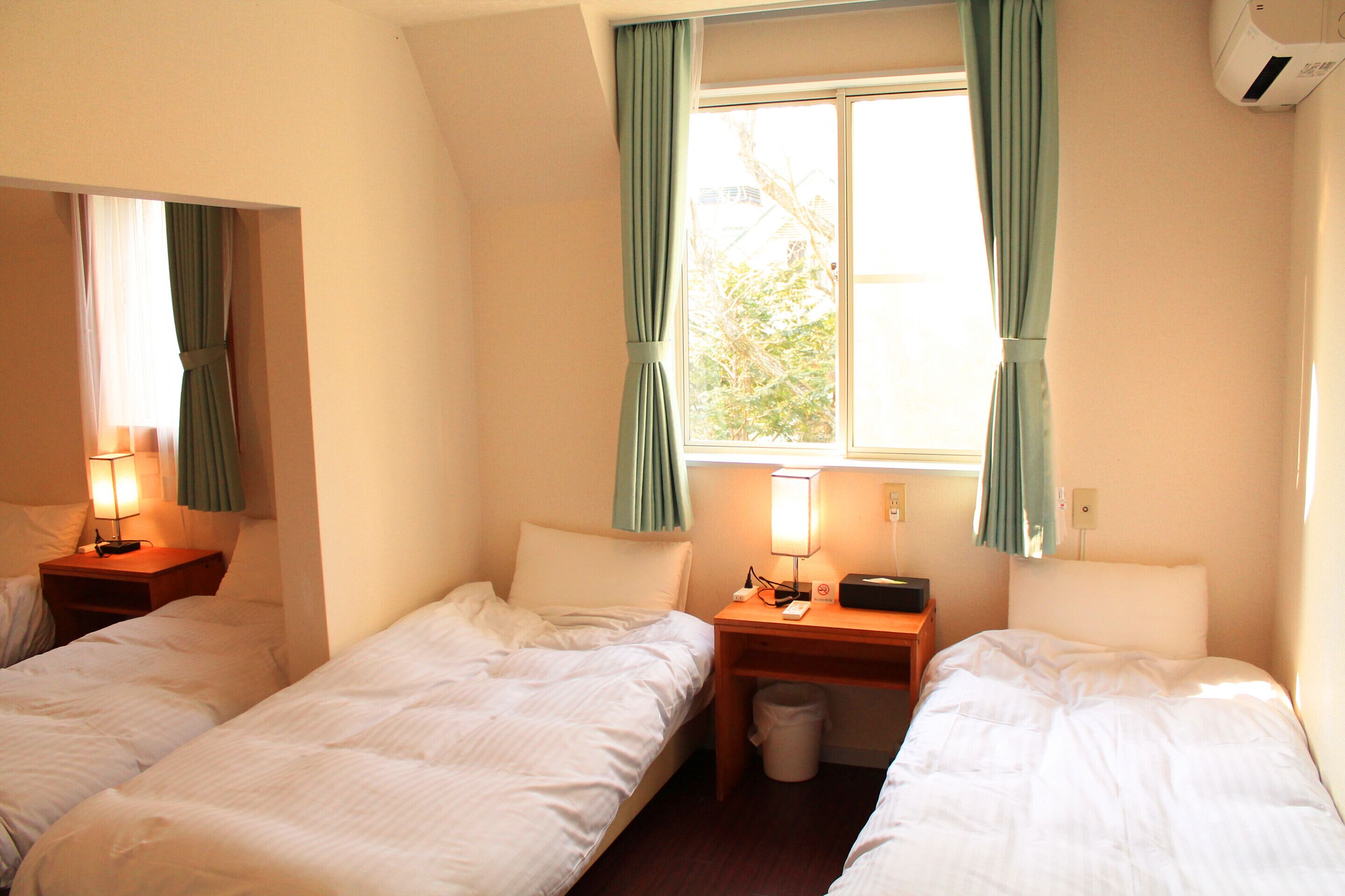 A hotel room with two single beds, a nightstand with a lamp, a small table with a phone and a trash bin, and a window with curtains.