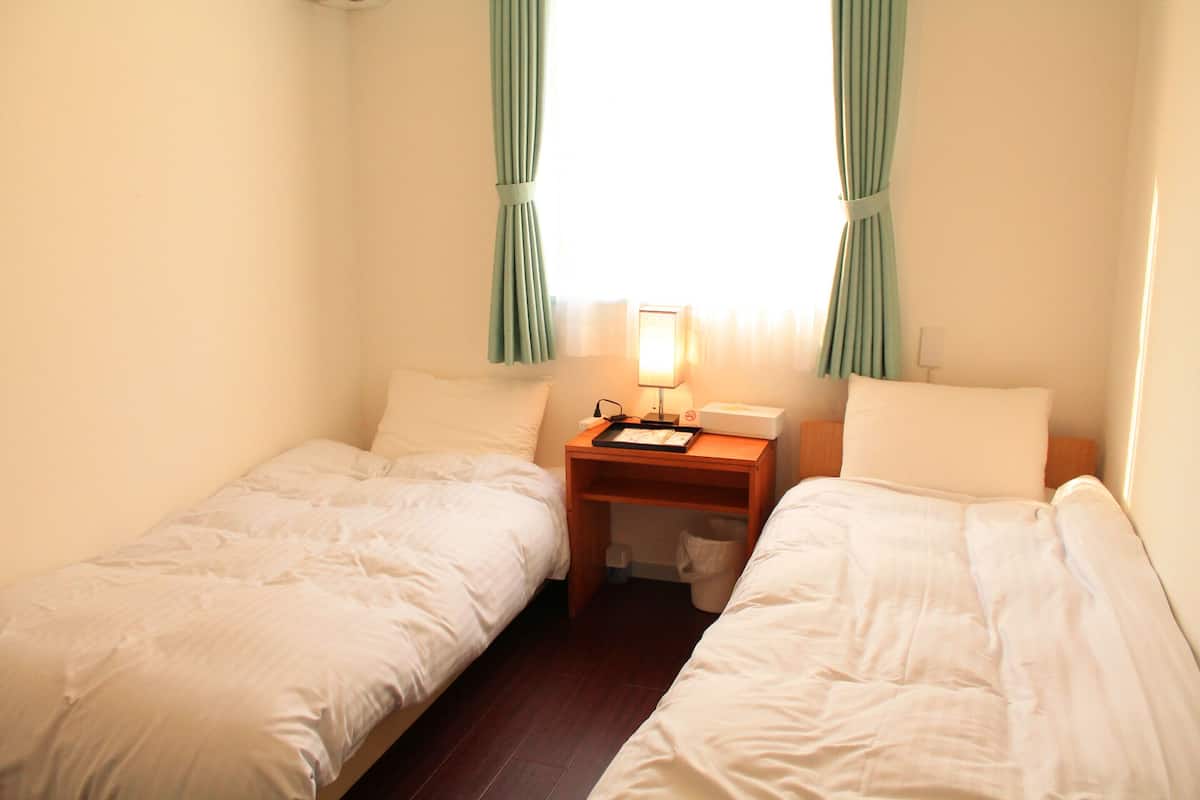 Quarto de hotel pequeno com duas camas de solteiro, uma mesinha de cabeceira com abajur e uma janela com cortinas.