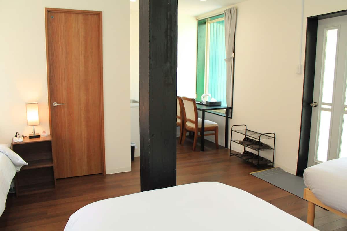 Quarto de hotel com porta de madeira, uma escrivaninha com cadeira, uma pequena mesa com abajur e um sapateiro.