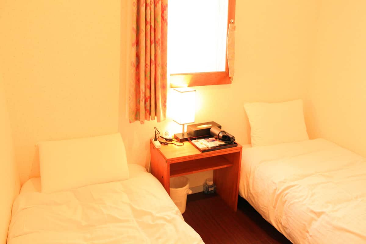 Quarto de hotel pequeno e individual, com duas camas, uma escrivaninha de madeira, uma luminária e uma janela com cortinas.