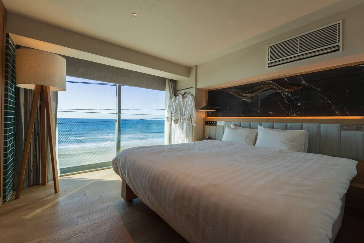 Una habitación de hotel con una cama grande, vista al océano, cabecera de mármol y un amplio ventanal.