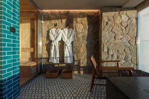 Un cuarto de spa con pared de piedra, bancos de madera y dos albornoces blancos colgados en ganchos.
