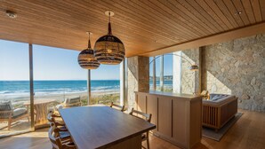 Een modern interieur met houten plafond, grote ramen met uitzicht op zee, een eethoek en een stenen muur.
