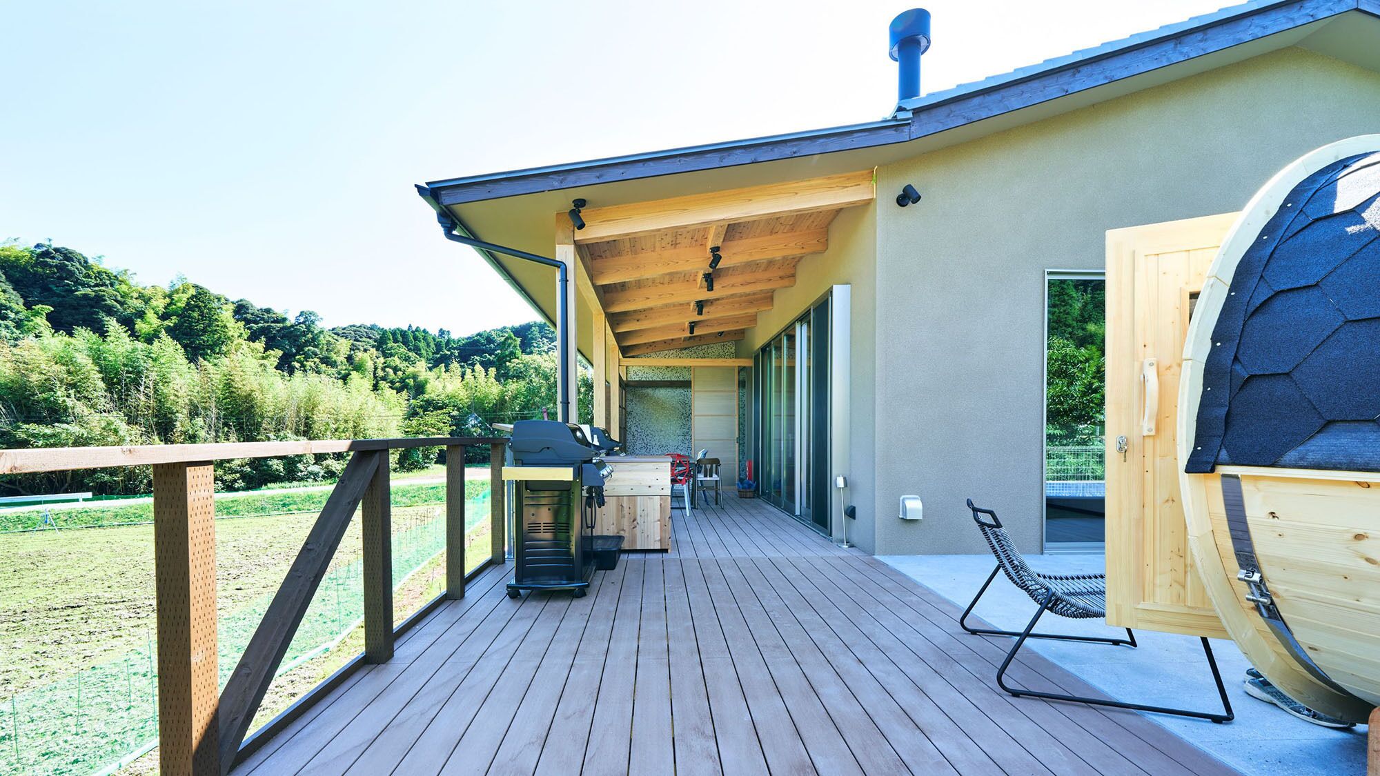 Una terraza de madera con una parrilla, una sauna y vistas a un área boscosa.