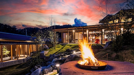 A modern house with a fire pit in the foreground, a waterfall, and a glass-enclosed dining area.