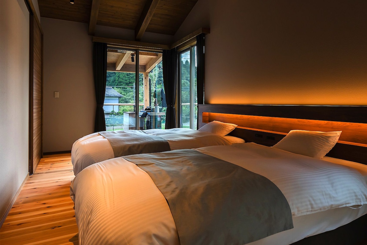 A bedroom with two beds, wooden flooring, a large window, and a view of greenery.