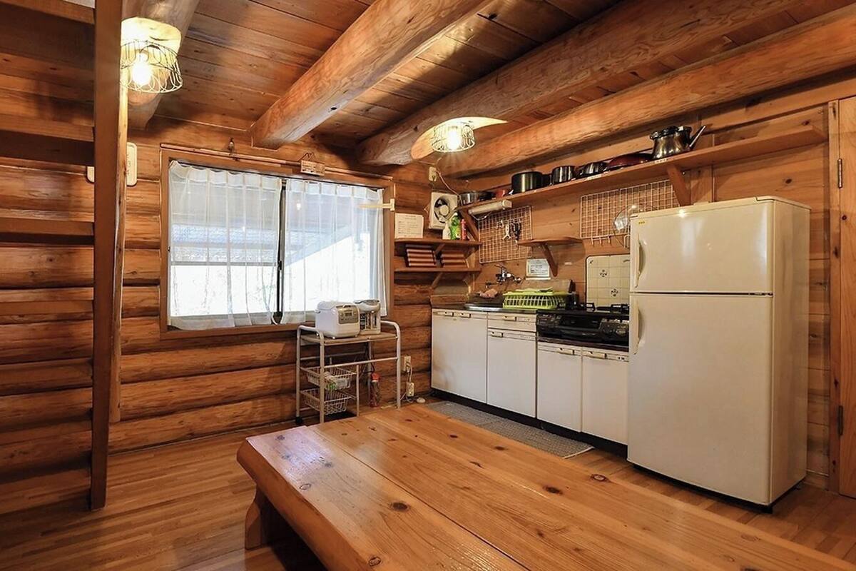 A log cabin interior with a wooden dining table, a kitchen with a refrigerator, and a window with curtains.