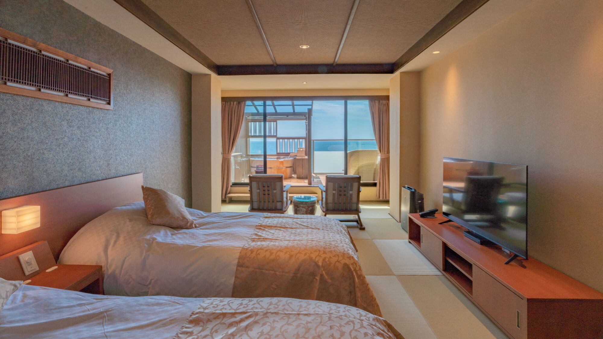 A hotel room with two beds, a TV, a seating area with a view of the sea, and a balcony.