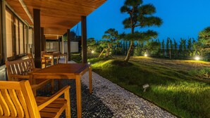 Un'area salotto esterna in legno con tavolo e sedie, circondata da un vialetto ghiaioso e un'area verde alberata.
