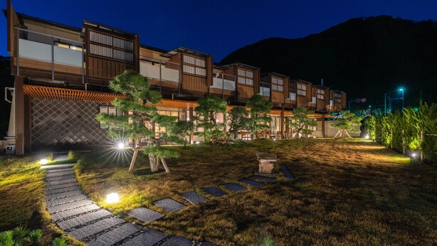 Un edificio moderno con diversi livelli, un giardino curato con sentieri in pietra e uno sfondo montuoso.