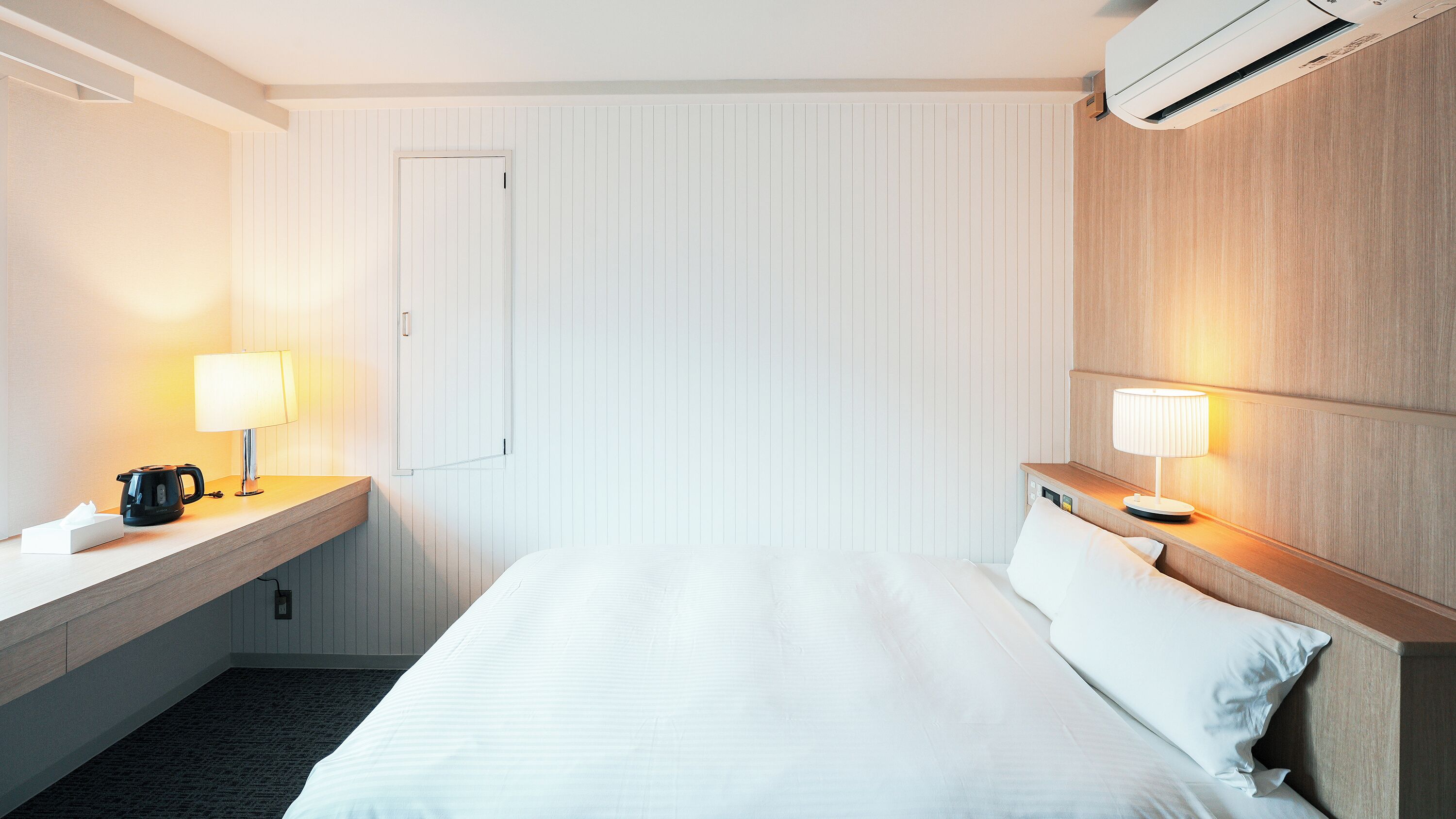 A hotel room with a bed, two bedside tables with lamps, a coffee maker, a tissue box, and an air conditioning unit.