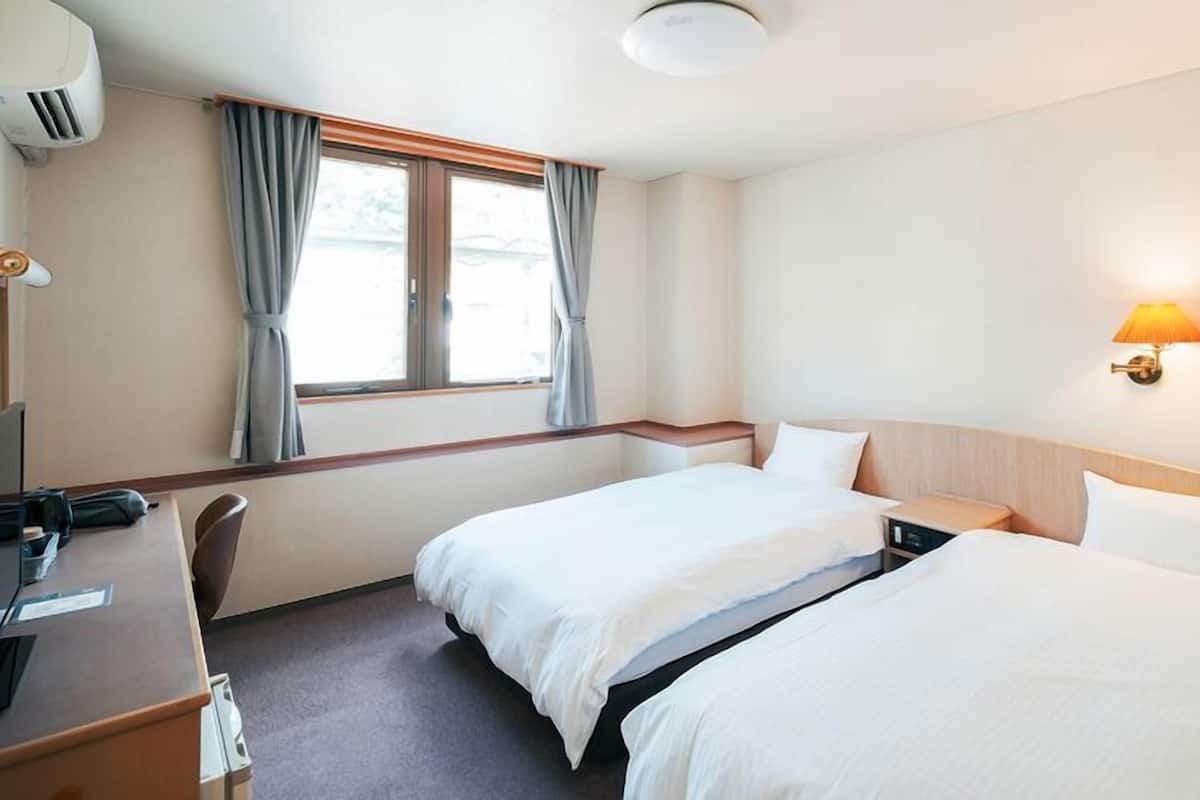 Ein Hotelzimmer mit zwei Betten, einem Schreibtisch mit Stuhl, einer Lampe und einem Fenster mit Vorhängen.
