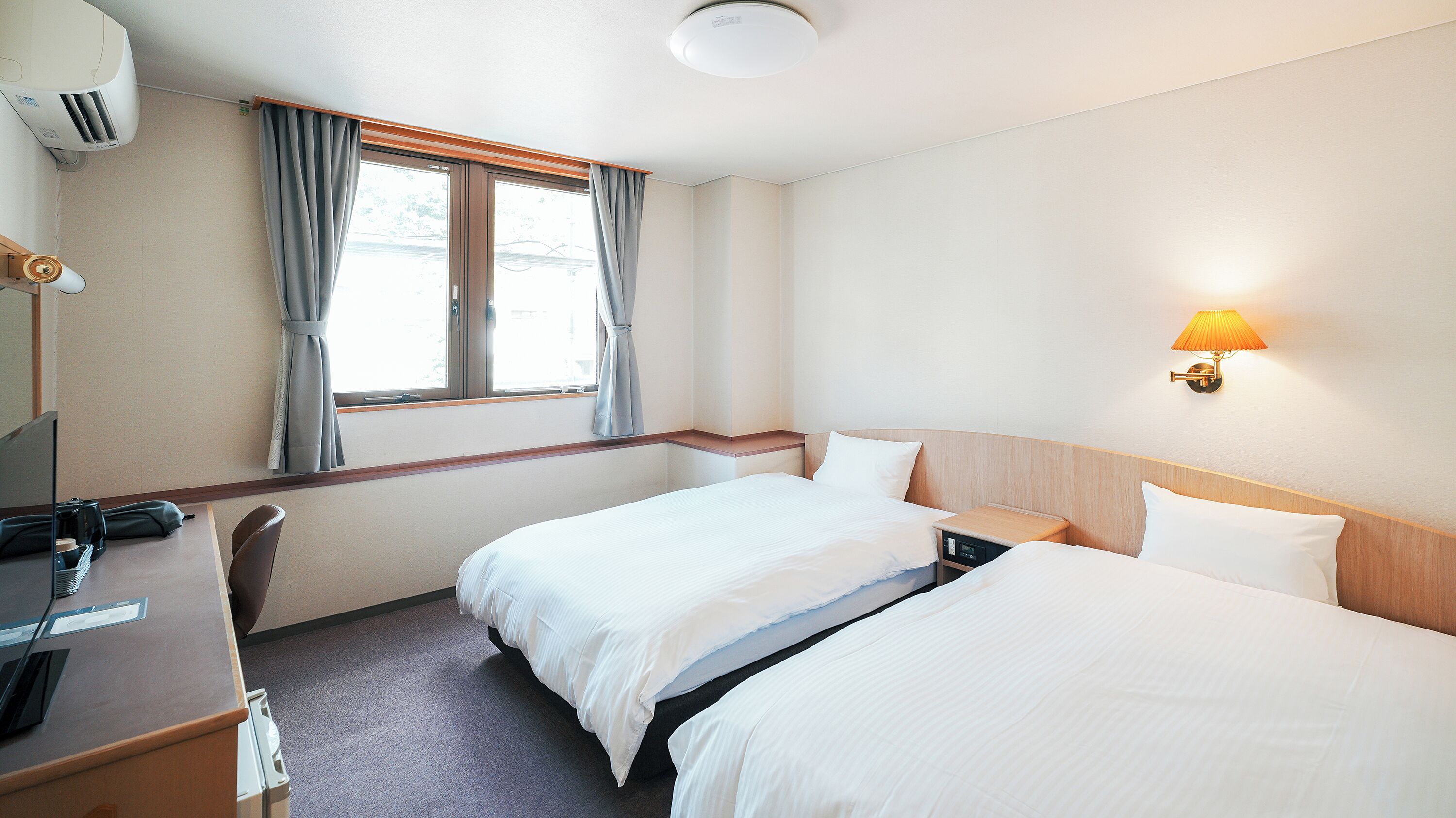 A hotel room with two beds, a desk with a computer, a lamp, and a window with curtains.