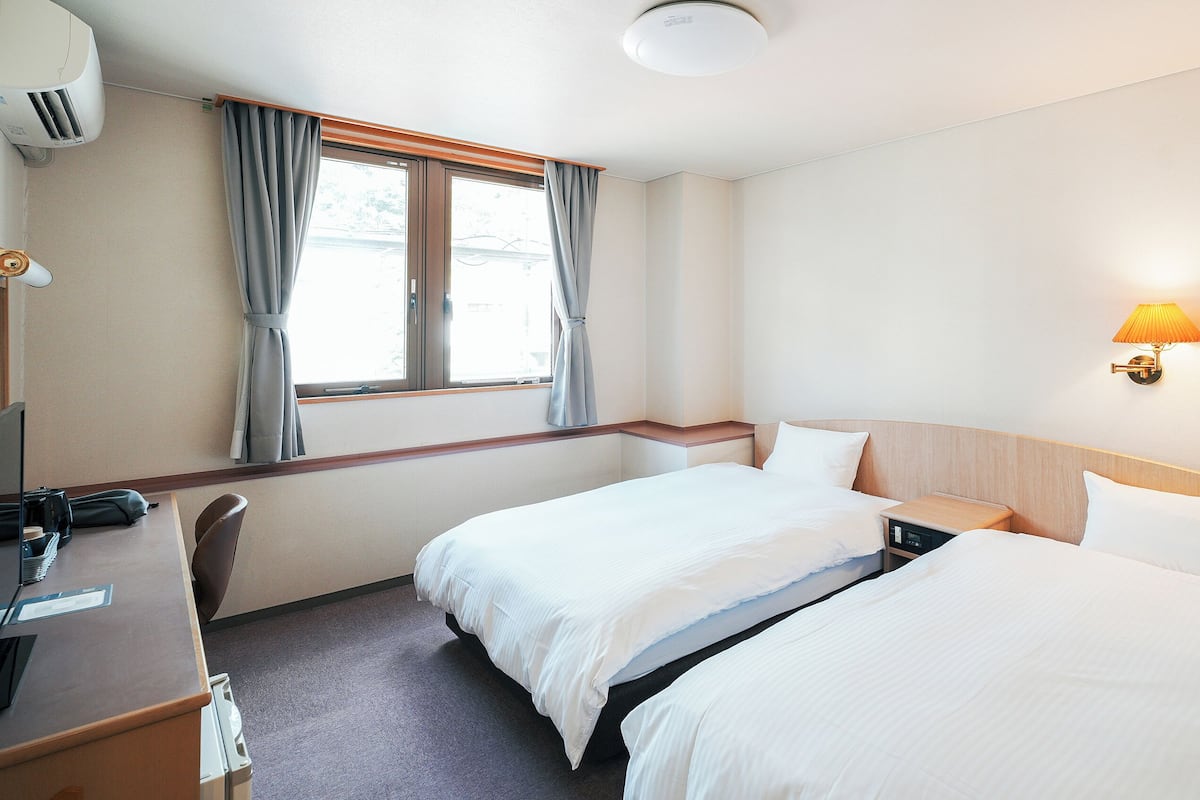 Ein Hotelzimmer mit zwei Betten, einem Schreibtisch mit Computer, einer Lampe und einem Fenster mit Vorhängen.