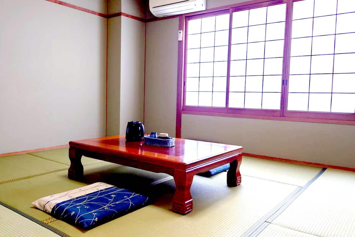 Ein Raum mit Tatami-Matten, einem niedrigen Holztisch, einem Kissen und einem Fenster mit Gitterläden.