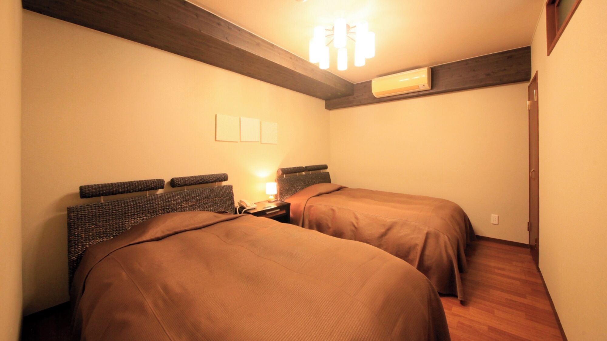 A room with two beds, a nightstand with a lamp, an air conditioner, and a wooden ceiling beam.