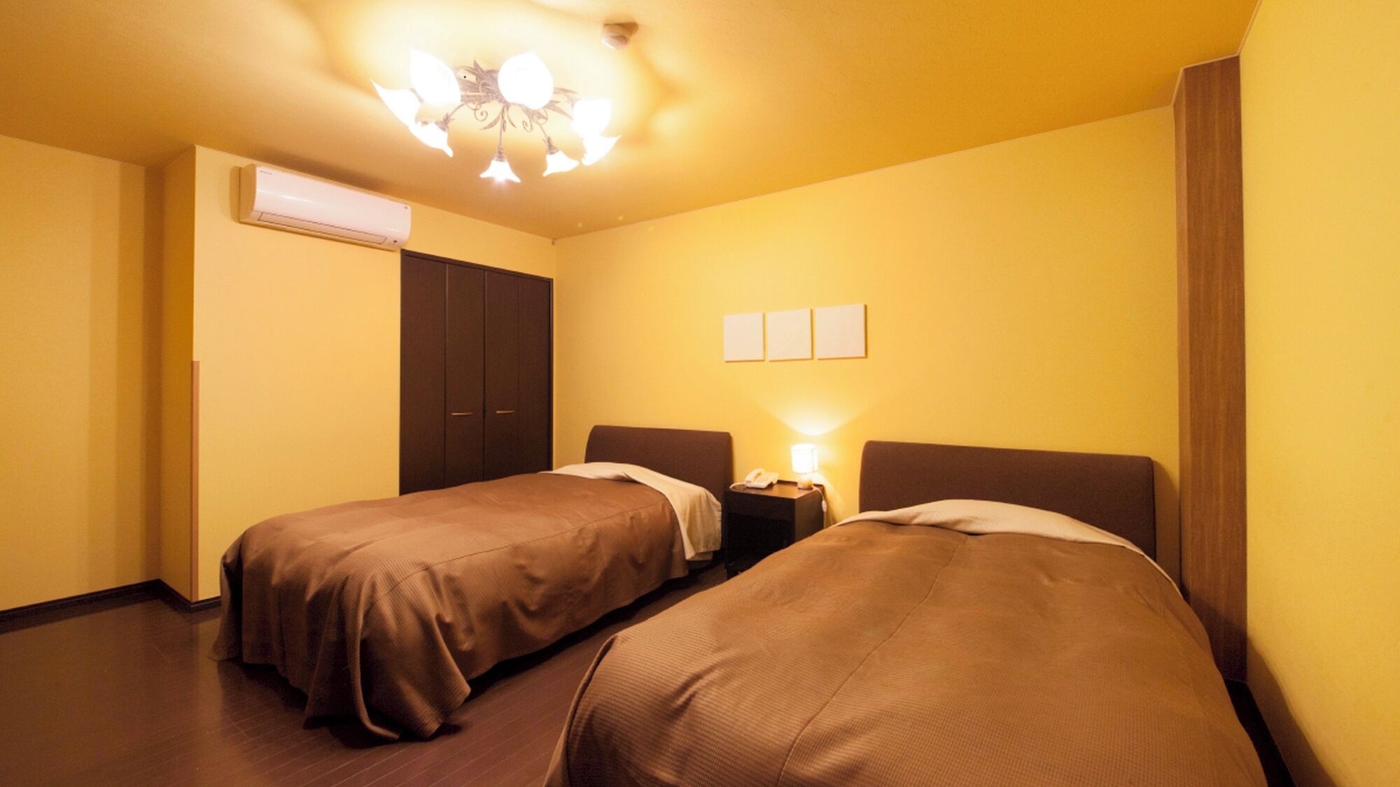 A hotel room with two beds, a wardrobe, an air conditioning unit, and a wall-mounted light fixture.