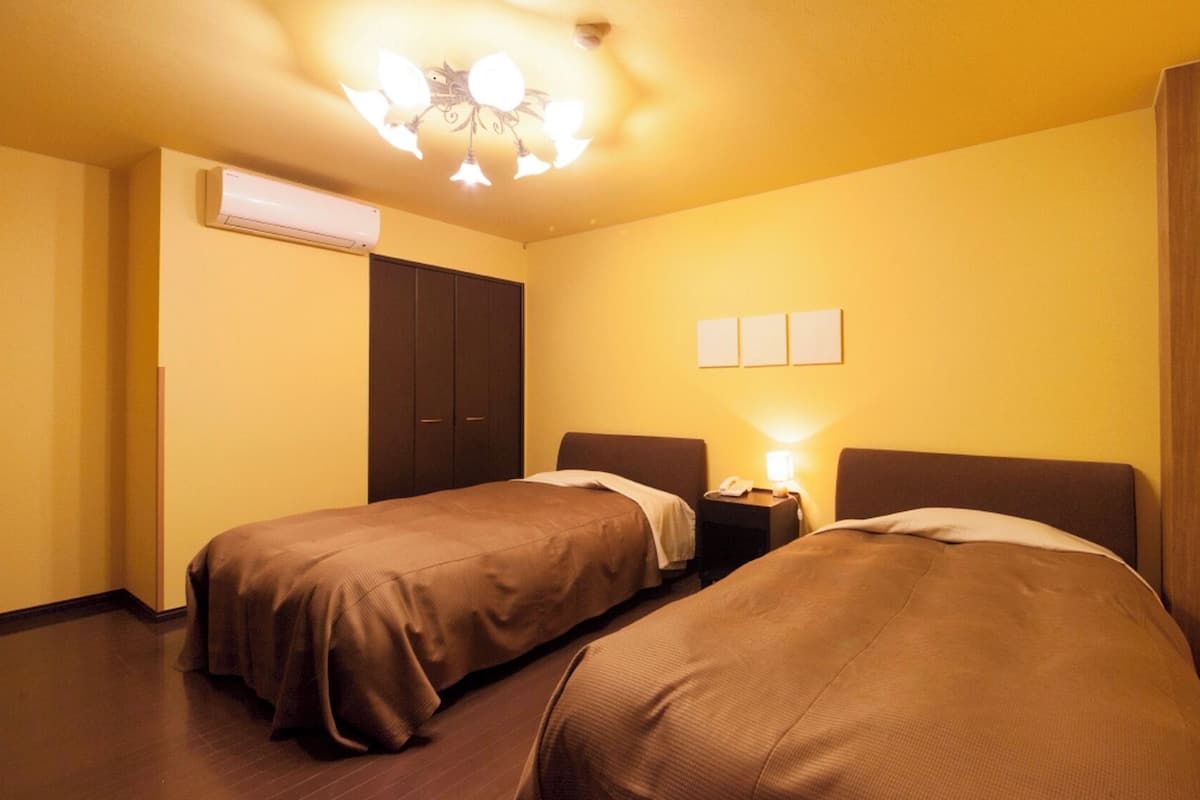 A hotel room with two beds, a wardrobe, an air conditioning unit, and a wall-mounted light fixture.