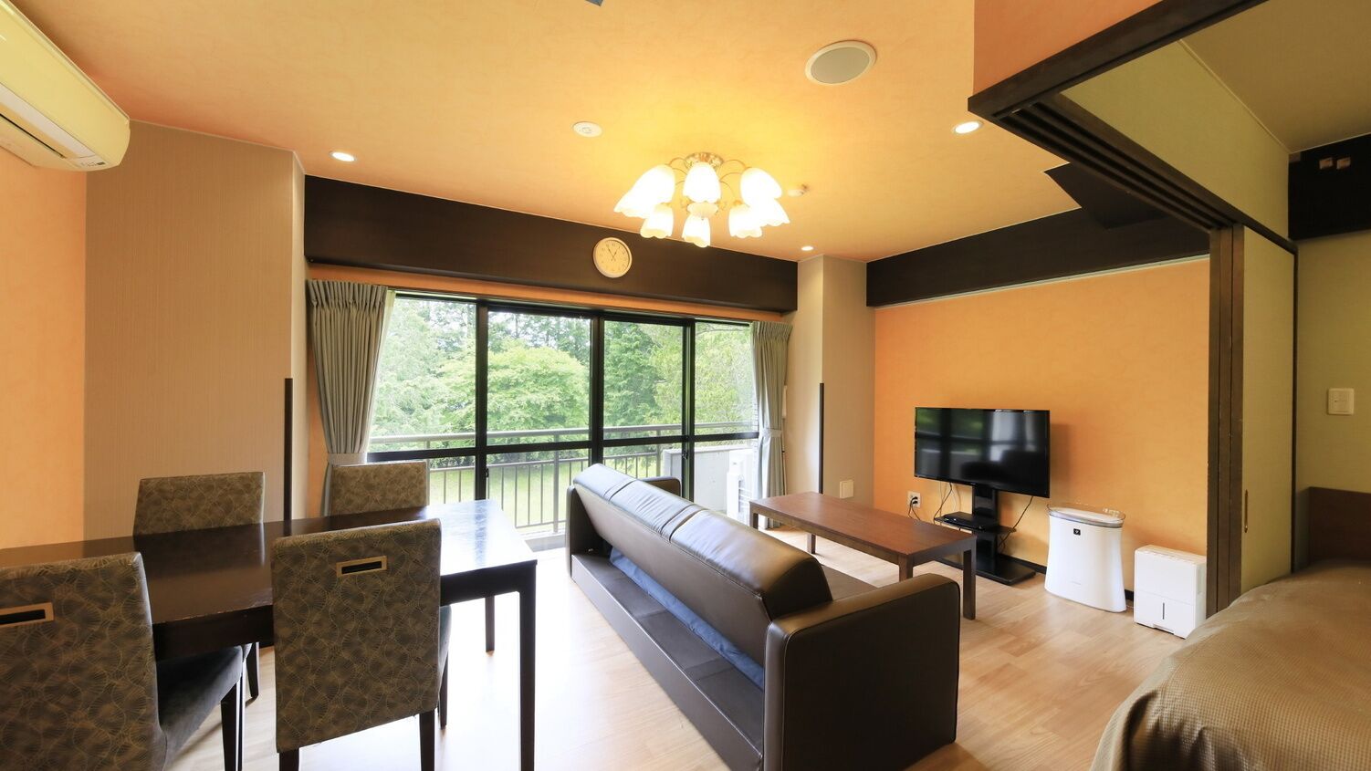 A hotel room with a dining area, a sofa, a television, and a window with a view of greenery.