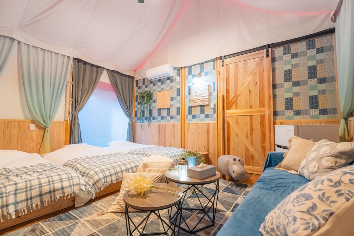 A room with two beds, a small table, a sofa, a wooden wardrobe, and a window.