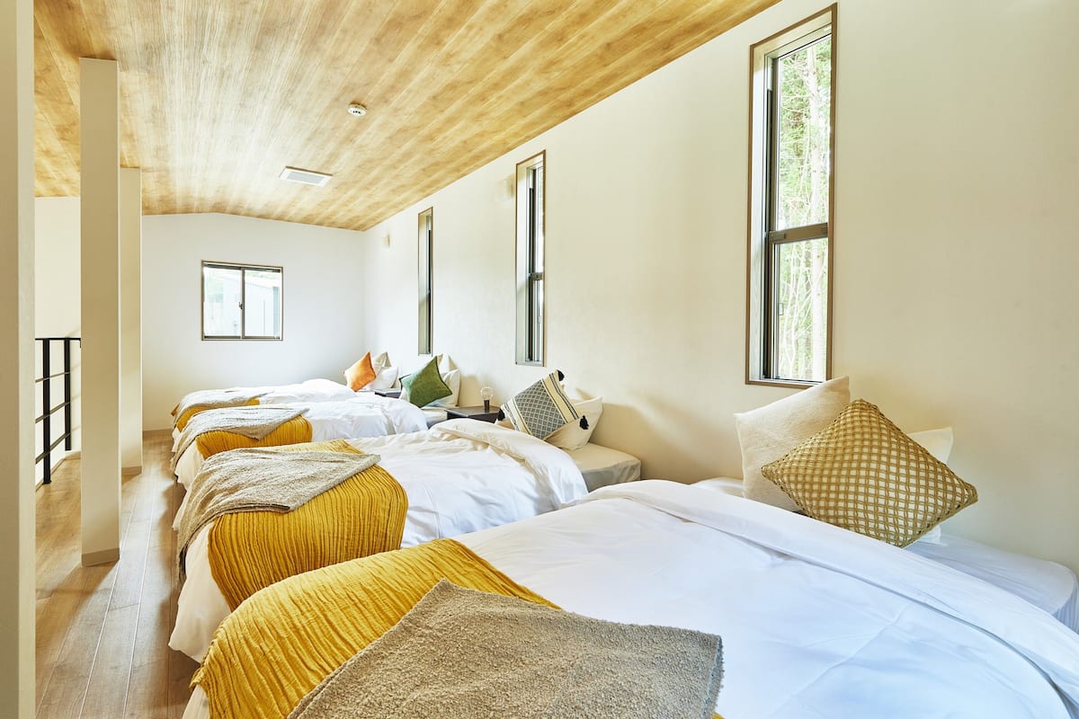 A hotel room with three beds, wooden ceiling, large windows, and a view of trees.
