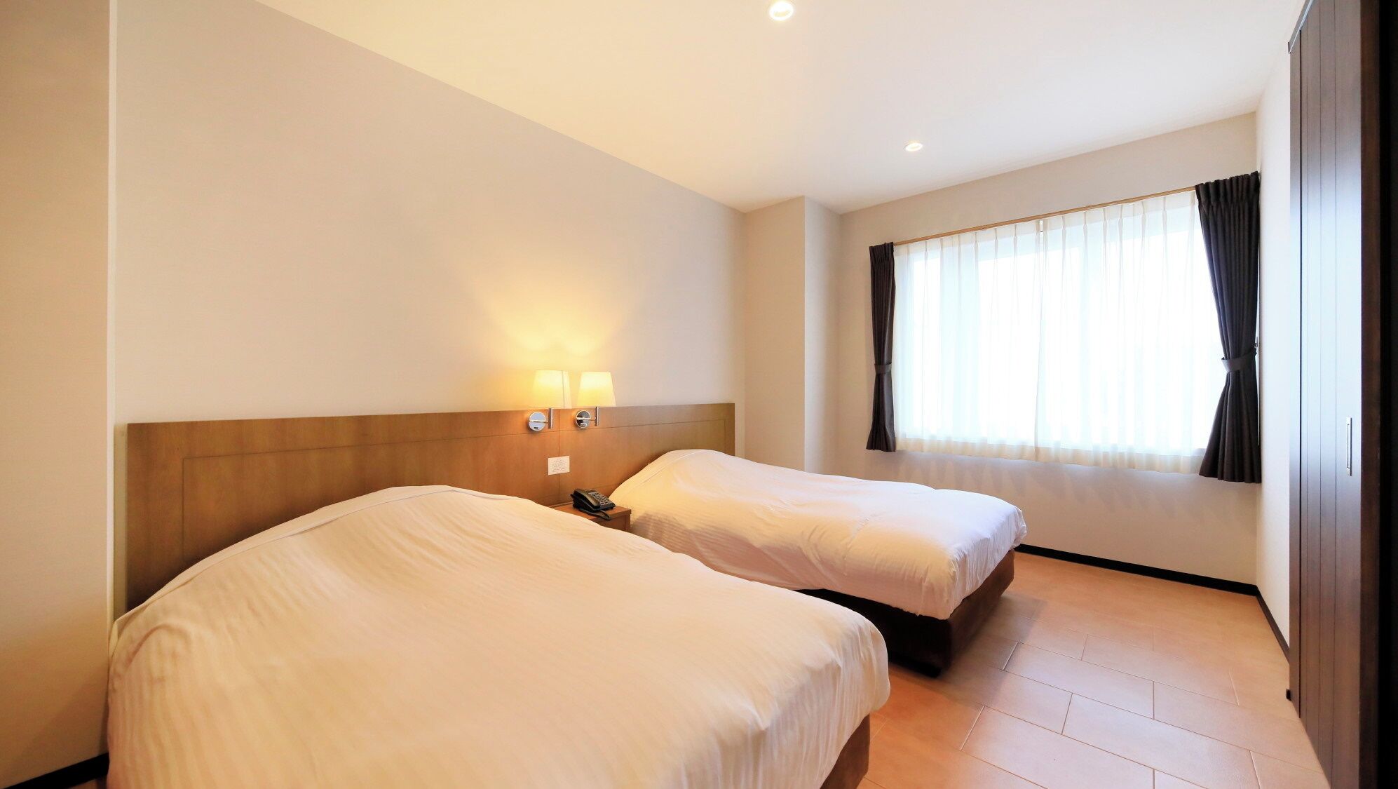 A hotel room with two beds, a nightstand with a phone, a wall-mounted lamp, and a window with curtains.