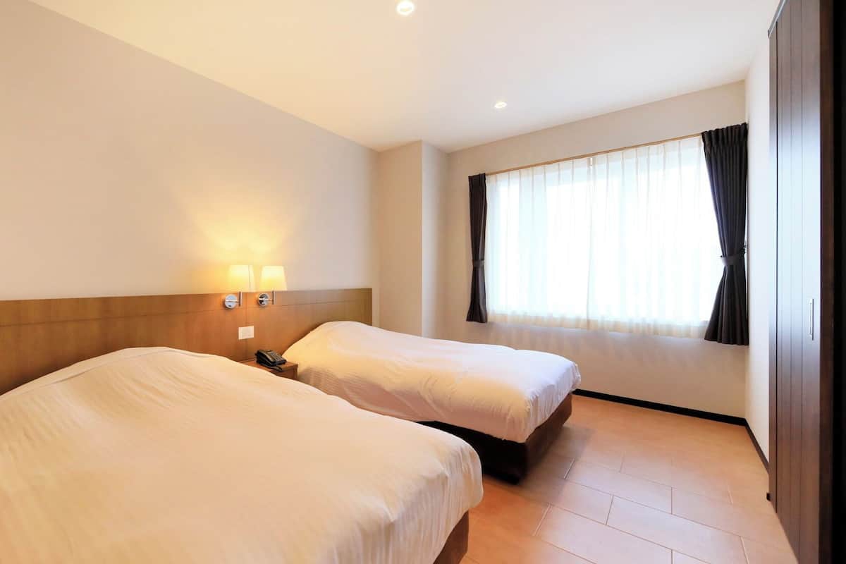 Une chambre d’hôtel avec deux lits, une table de chevet avec un téléphone, une lampe fixée au mur et une fenêtre avec des rideaux.
