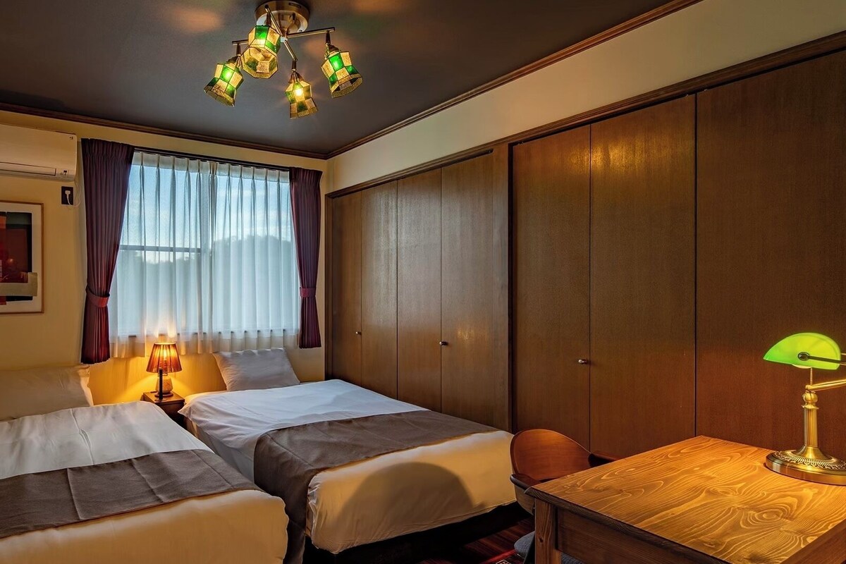 Une chambre d’hôtel avec deux lits, un bureau, une lampe, un luminaire au plafond et une fenêtre avec des rideaux.