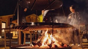 Un foyer extérieur circulaire, doté d’une grille en métal, entouré d’une table et de chaises en bois.