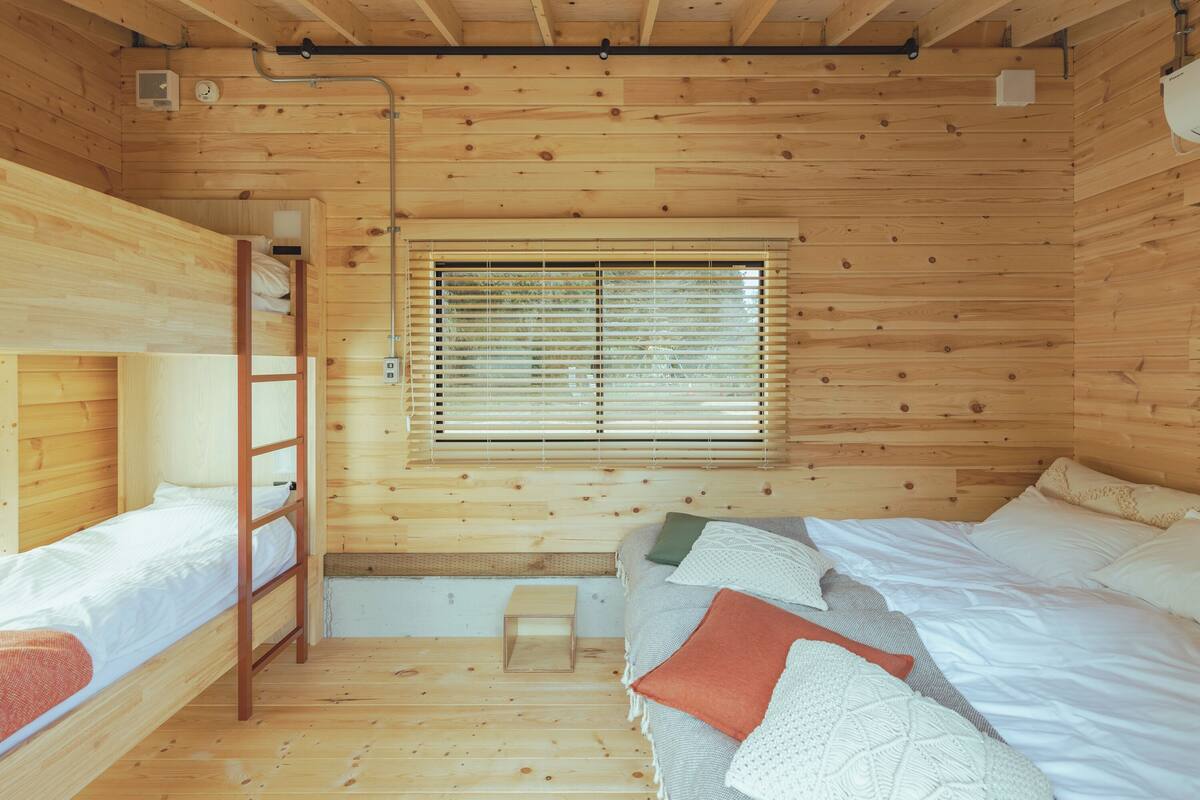 Een houten kamer met stapelbedden, een ladder, een raam met jaloezieën en een klein krukje.