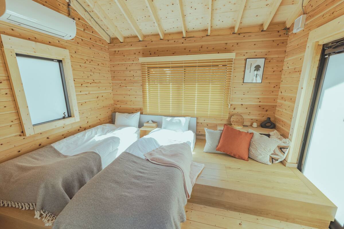 Een houten kamer met twee bedden, een raam met zonwering, een klein tafeltje en een deur.