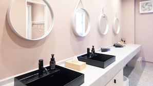 Une salle de bain avec trois lavabos, chacun doté d’un receveur noir et d’un miroir au-dessus. On peut voir des articles de toilette sur le comptoir, et un porte-serviettes est visible dans le reflet du miroir.
