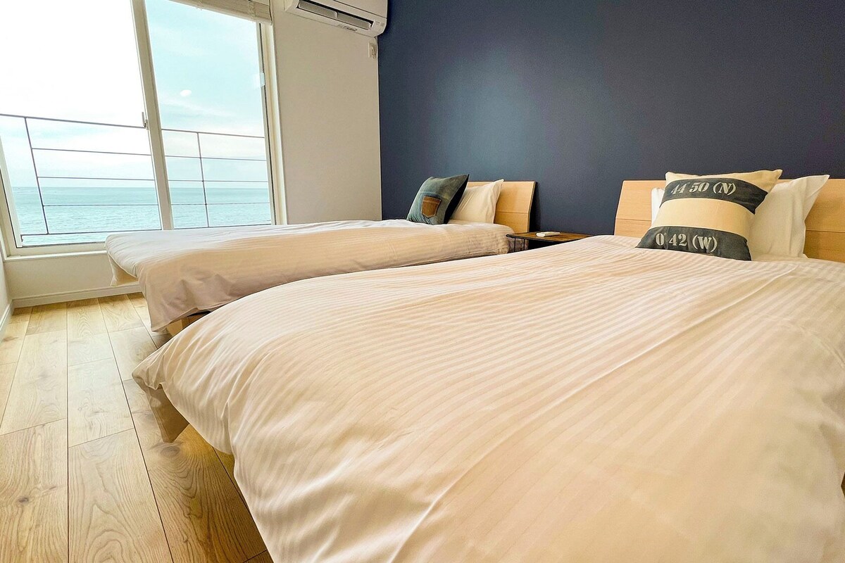 Une chambre avec deux lits, une vue sur la mer et un mur bleu foncé.
