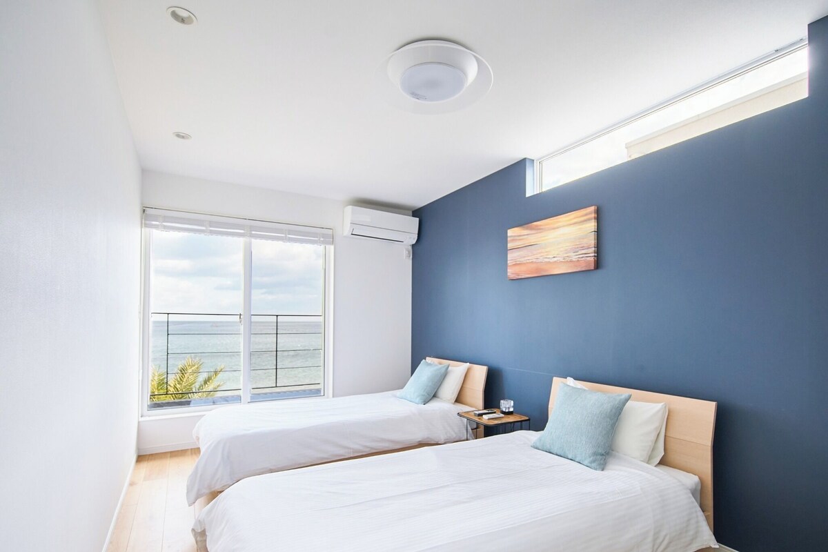 Une chambre avec deux lits, une grande fenêtre donnant sur la mer et un luminaire au plafond.