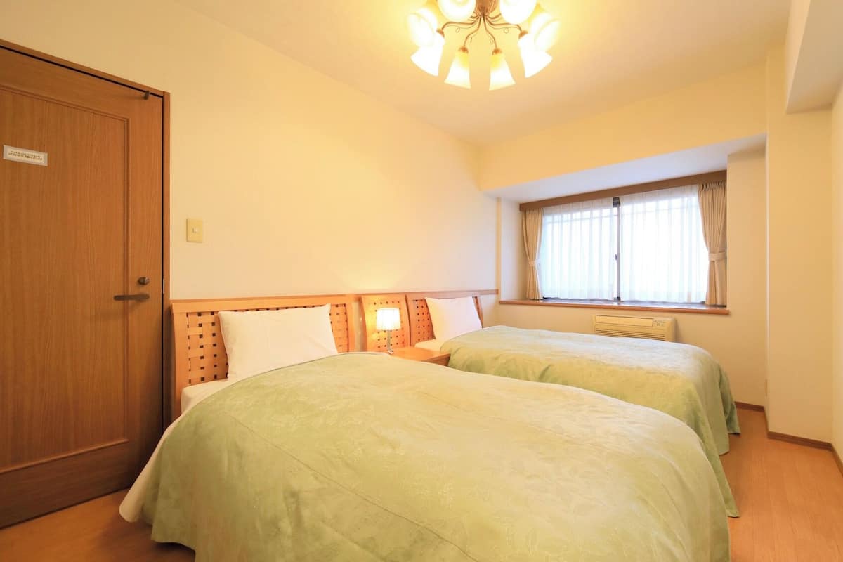 Ein Hotelzimmer mit zwei Betten, einer Holztür, einem Fenster mit Vorhängen und einer Deckenleuchte.