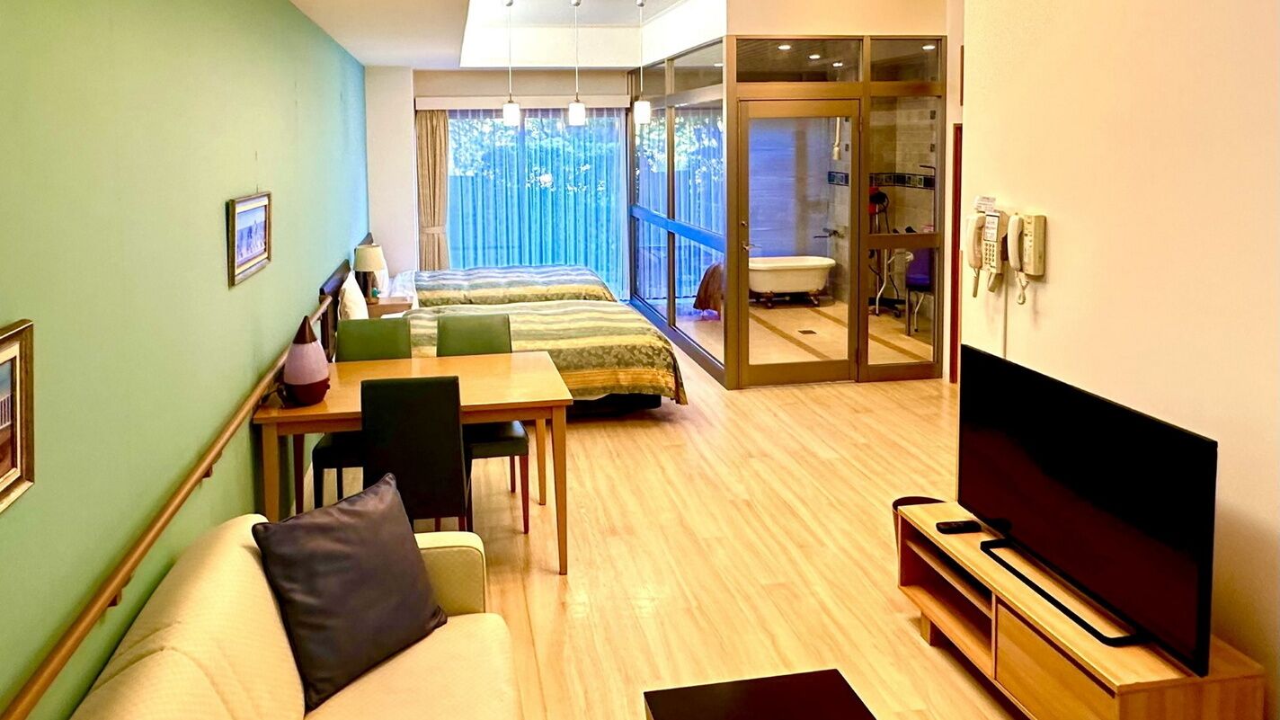 A hotel room with a bed, a TV, a desk, and a bathroom visible through a glass door.