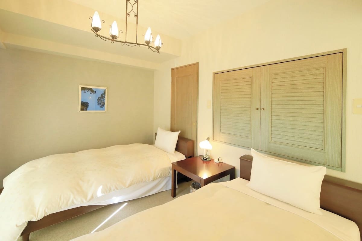 Ein Hotelzimmer mit zwei Betten, einem Nachttisch, einer Lampe, einem gerahmten Bild an der Wand und einem großen Fenster mit Fensterläden.