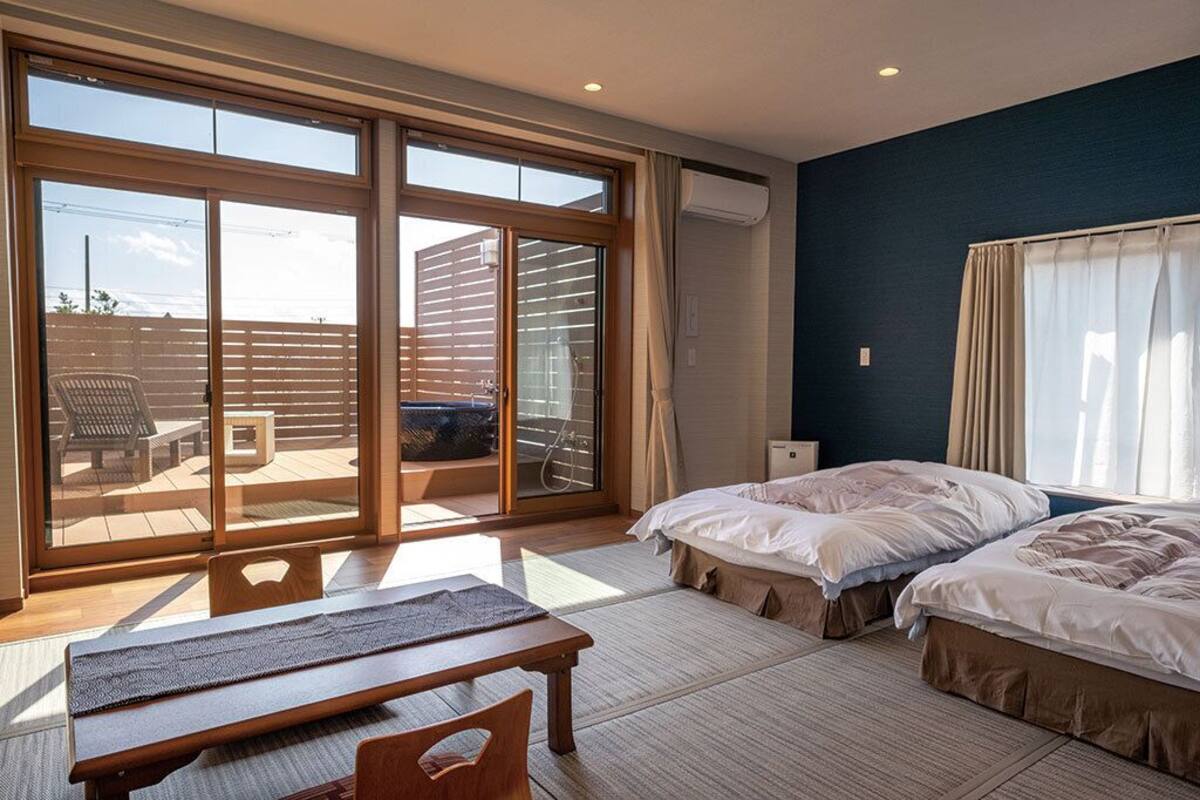 Une chambre d'hôtel avec deux lits, un banc, une table et de grandes portes-fenêtres coulissantes donnant sur un espace extérieur.
