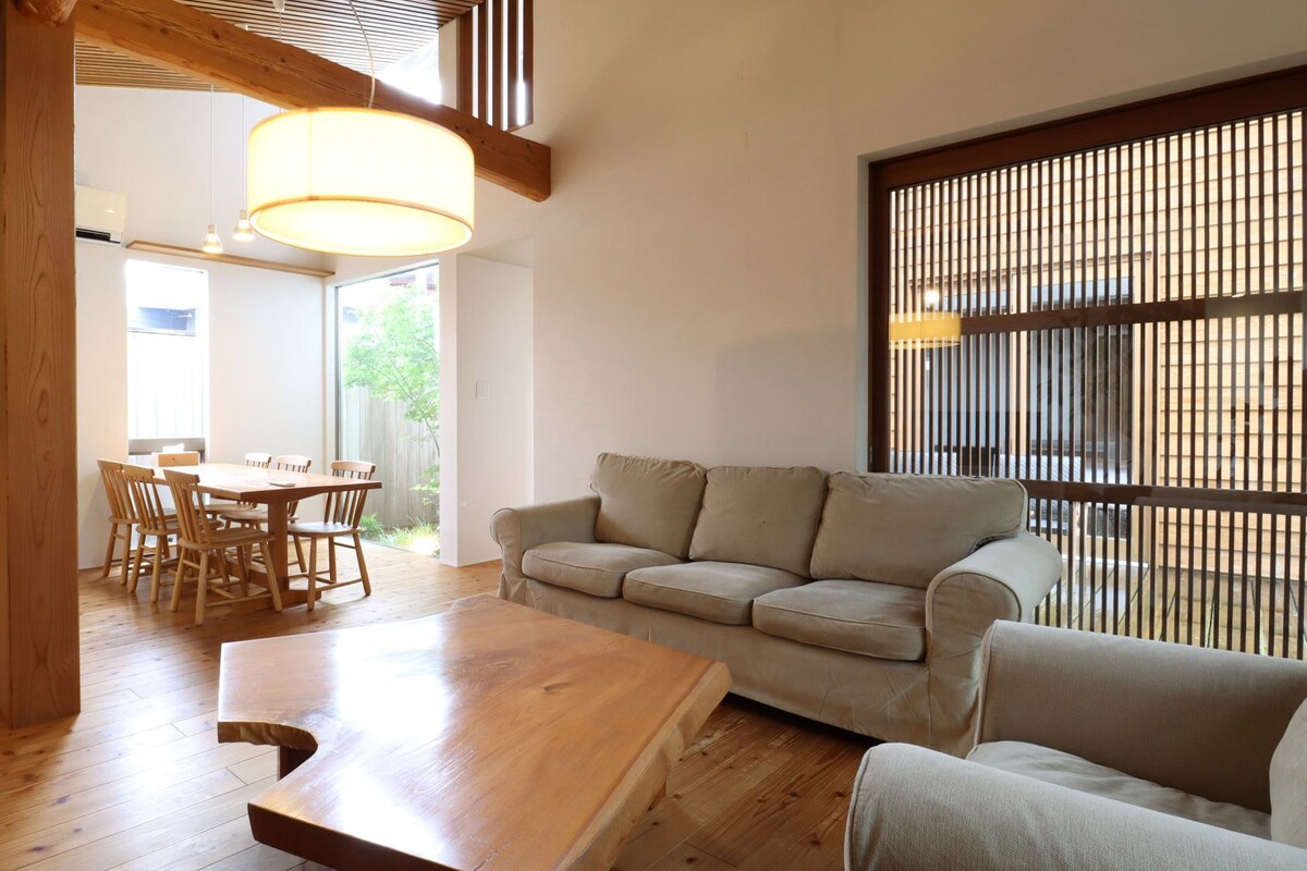 A modern living room with a wooden dining table, a sofa, and a large window with vertical blinds.