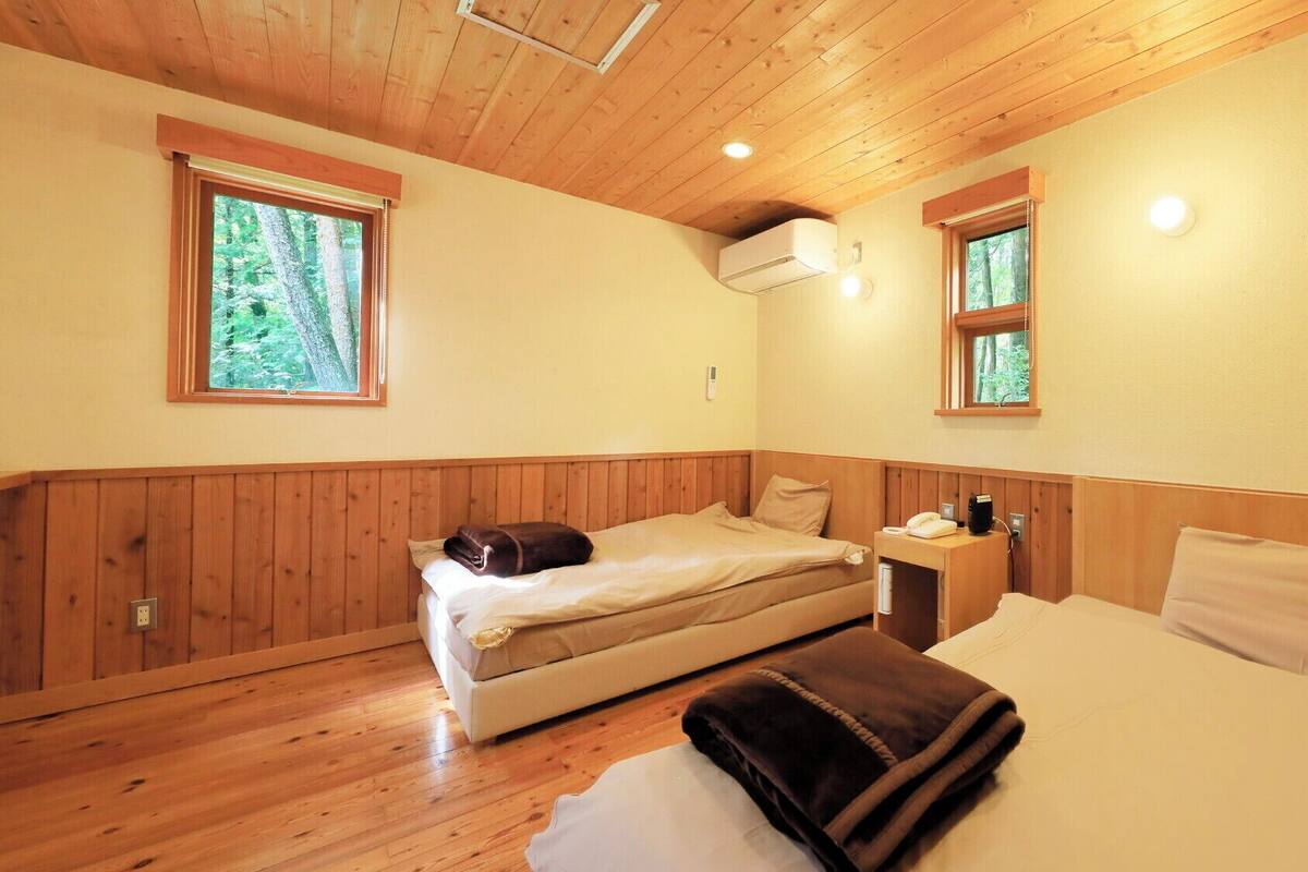 Un dormitorio con techo de madera, dos ventanas con vistas a zonas verdes, una cama con manta, una mesita de noche con teléfono y un equipo de aire acondicionado.