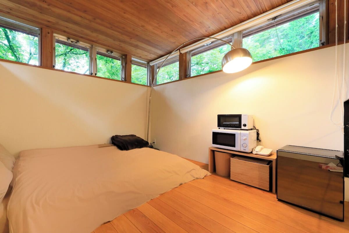 Un dormitorio con cama, mesita de noche con microondas y teléfono, escritorio con computadora y ventanas amplias.