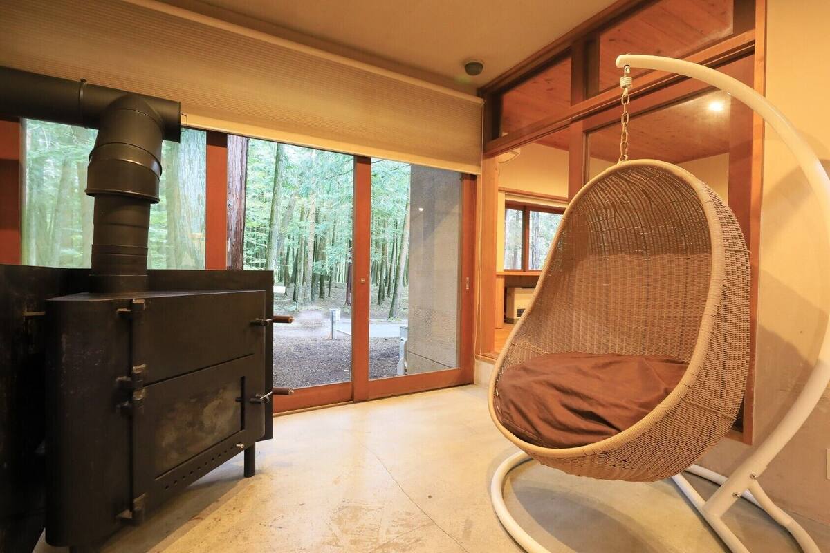 Una habitación con estufa de leña, una silla colgante y grandes ventanales con vistas al bosque.