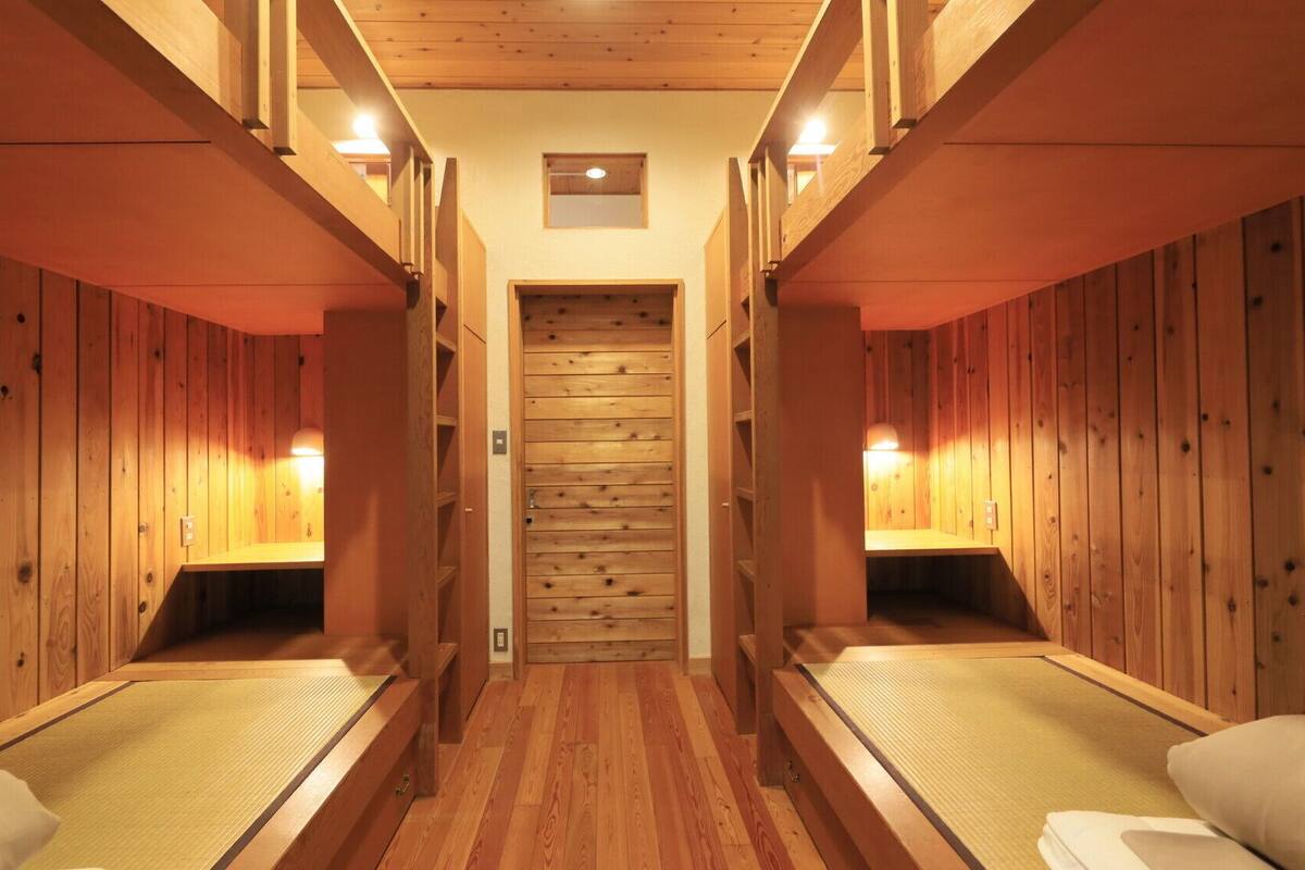 Una hilera de cabañas de madera con camas individuales, una puerta central y luces fijadas a la pared.